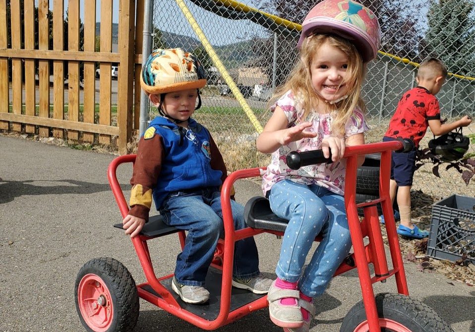 Girl and boy riding a tricycle while smiling.